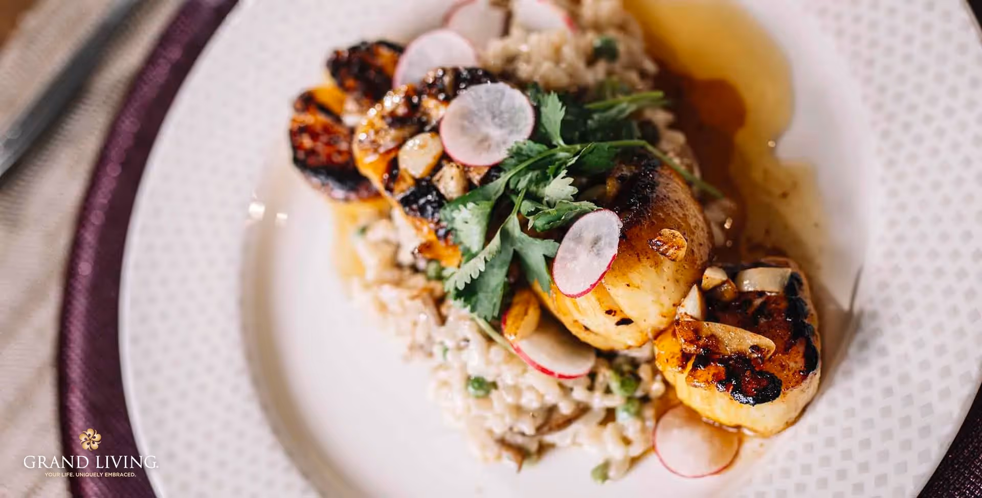Close-up of a plated scallop dish with risotto, radish slices, and greens on a patterned plate.