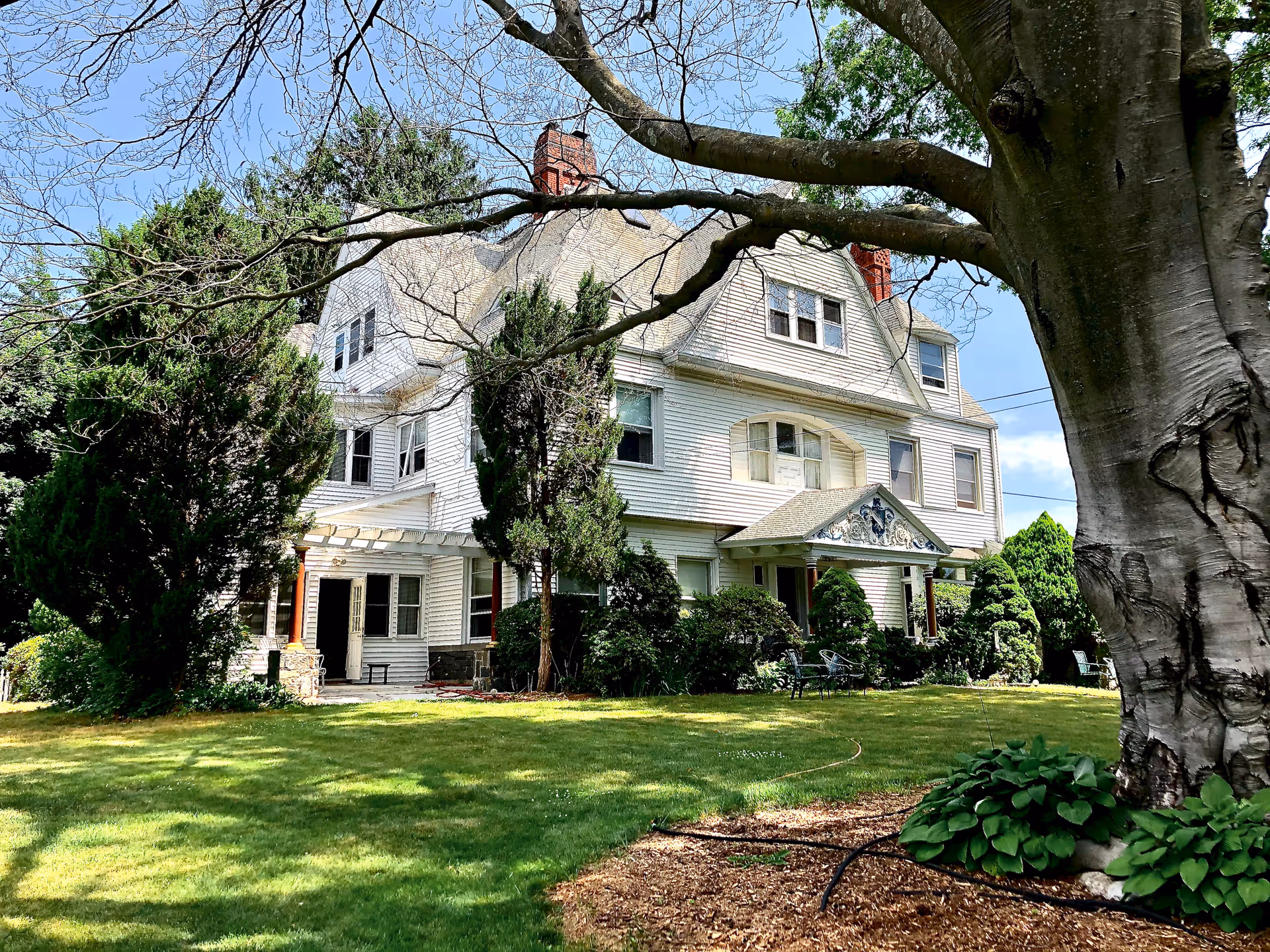 A large white multi-story house with a steep roof and multiple windows, surrounded by green trees and bushes. There is a well-maintained lawn in the foreground with a large tree on the right side of the image. The sky is clear and blue.