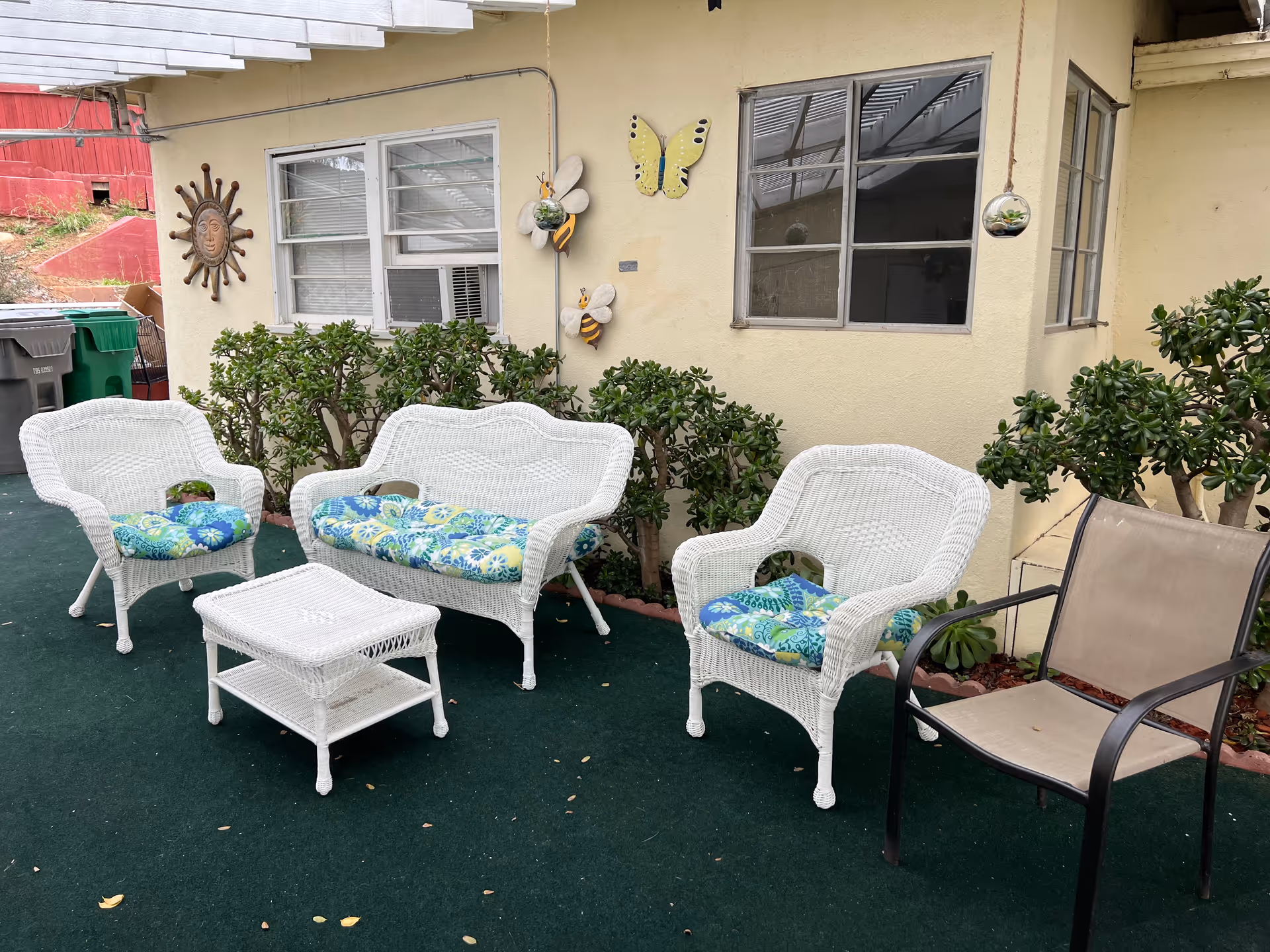 Outdoor patio area with white wicker furniture including two chairs, a loveseat, and a small table. The chairs and loveseat have blue and green patterned cushions. There is also a beige metal-framed chair. The patio is covered with a green outdoor carpet and is adjacent to a yellow building wall decorated with a sun sculpture, two bee decorations, and a butterfly decoration. There are green bushes along the wall and hanging glass terrariums.