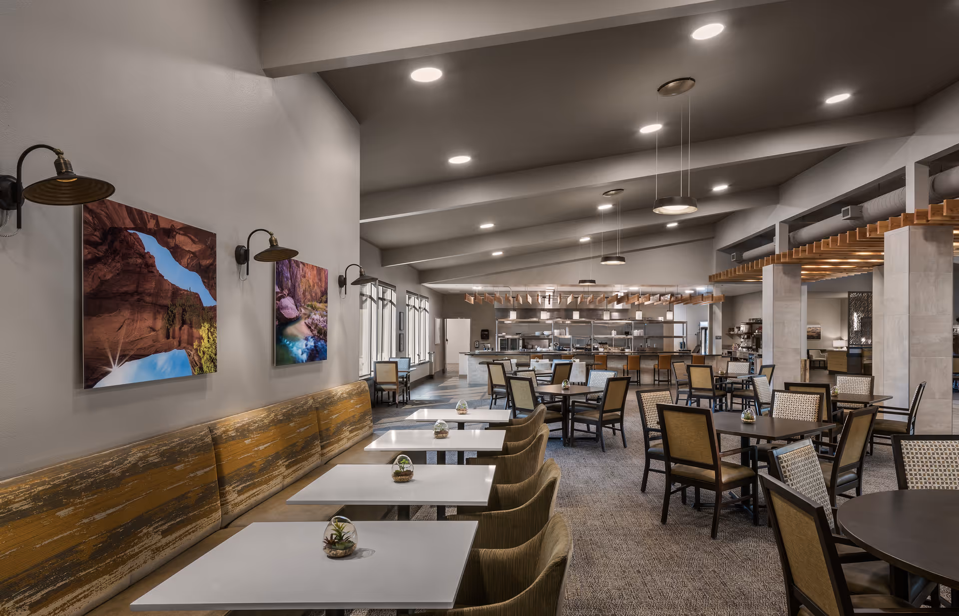 A spacious dining room with multiple tables and chairs arranged neatly. The room features a long bench with a rustic wooden backrest along one wall, decorated with landscape photographs and wall-mounted lamps. The ceiling has recessed lighting and hanging pendant lights. In the background, there is an open kitchen area with stainless steel appliances and a counter with bar stools.