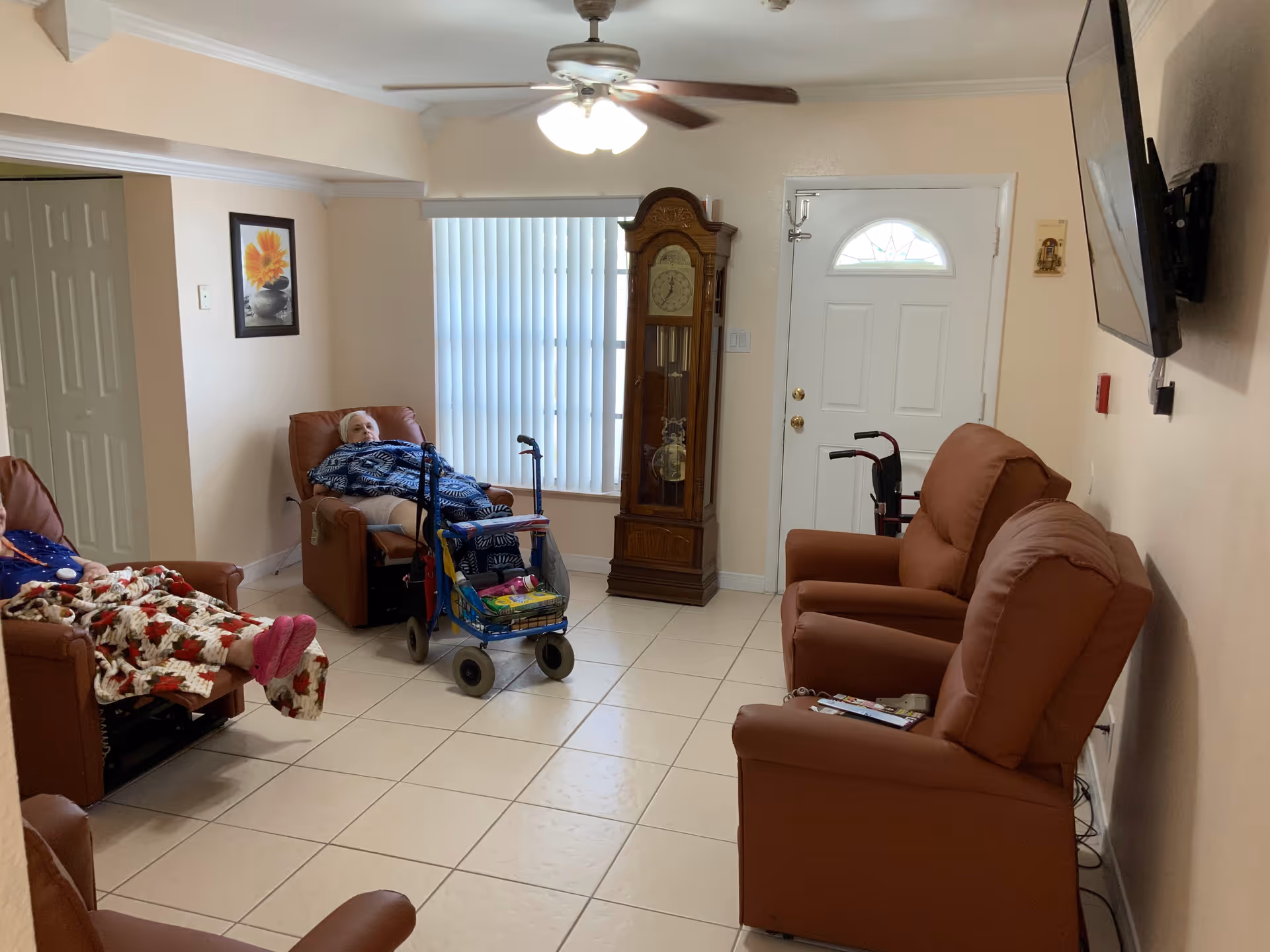 A small living room area with four brown recliner chairs arranged around the room. Two elderly women are seated in two of the chairs, each covered with a blanket. A walker with a basket is positioned near one of the women. There is a grandfather clock against the wall next to a window with vertical blinds. A ceiling fan with lights is mounted on the ceiling, and a flat-screen TV is mounted on the wall to the right. The room has tiled flooring and light-colored walls with a framed flower picture hanging on one wall.