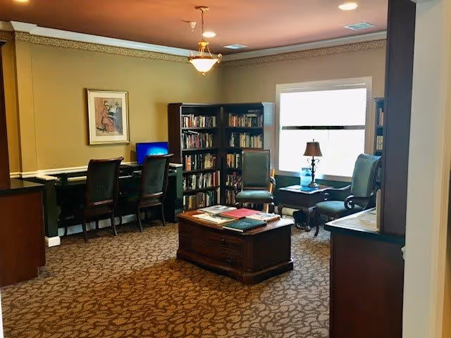 A cozy interior room with beige walls and patterned carpet featuring a wooden coffee table in the center with books on it. There are two green upholstered chairs near a window with a small table and lamp between them. A bookshelf filled with books is against the wall, and a desk with two chairs and a computer monitor is on the left side. A framed picture hangs on the wall above the desk, and a ceiling light fixture is visible.