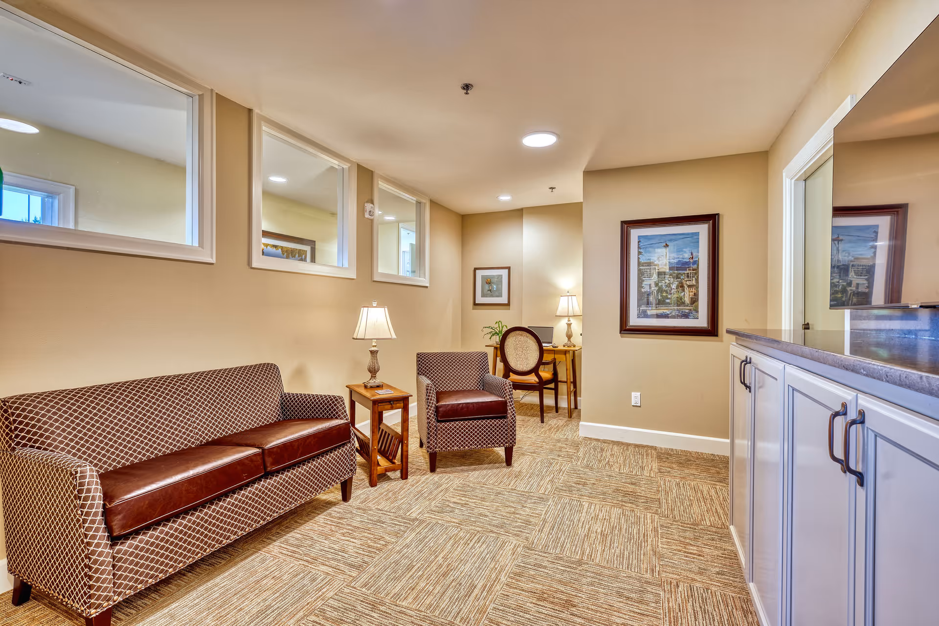 A cozy interior room with a patterned sofa and matching armchair, a wooden side table with a lamp, a small desk with a chair and lamp in the corner, framed artwork on the walls, and a cabinet with a countertop on the right side.