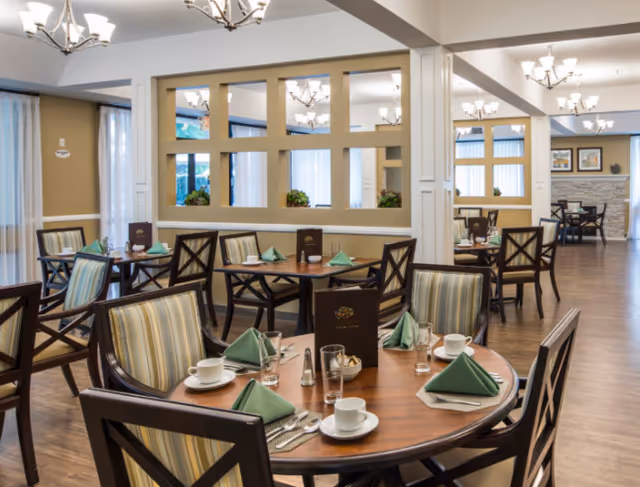 A bright and spacious dining room with multiple round wooden tables set with green folded napkins, white cups and saucers, glasses, and silverware. The chairs have striped upholstery and dark wooden frames. The room features large windows with vertical blinds, beige walls, and multiple chandeliers providing warm lighting. A decorative partition with square openings separates different sections of the room.