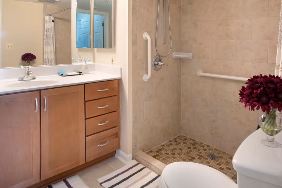 A bathroom with a wooden vanity featuring a white countertop and sink, a large mirror above it, a walk-in tiled shower with grab bars and a handheld showerhead, a white toilet with a vase of dark purple flowers on top, and a striped bath mat on the floor.