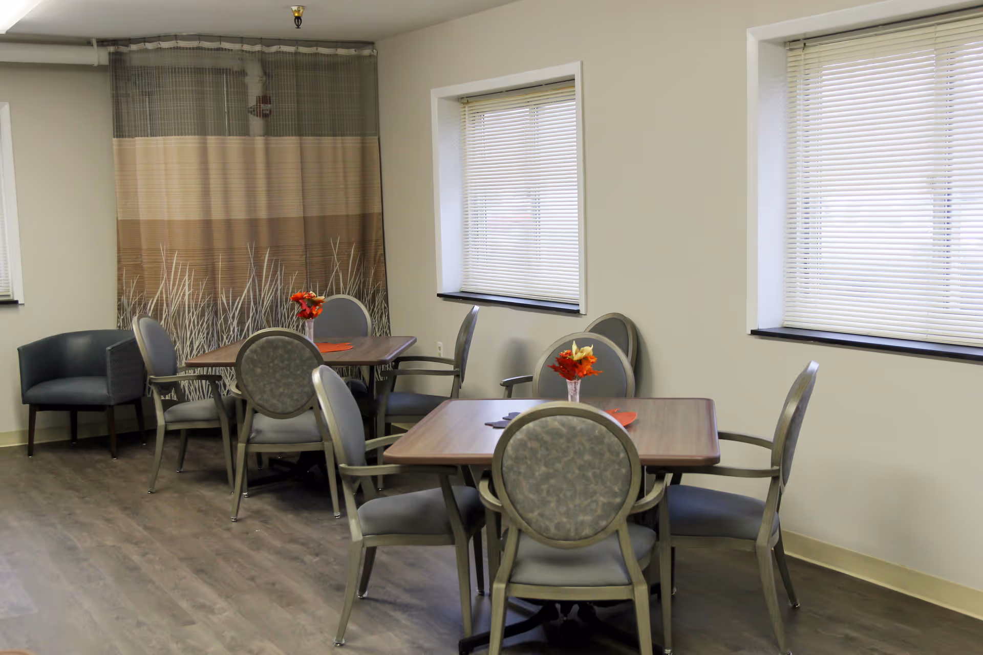 A small dining area with two square wooden tables, each surrounded by four cushioned chairs with armrests. Each table has a small vase with red and orange flowers. The room has light-colored walls, two windows with blinds, a patterned curtain on one wall, and a single dark armchair in the corner.