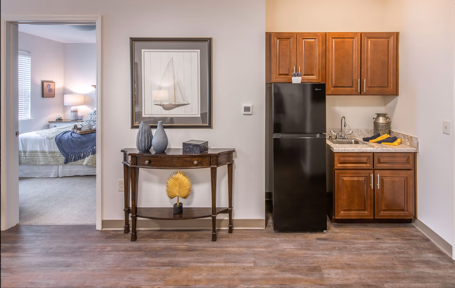 Small interior featuring a kitchenette with a black refrigerator and wood cabinets, a decorative console table, and a doorway opening to a bedroom.
