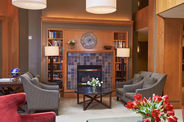A cozy living room area with a tiled fireplace centered on a wooden wall unit that includes bookshelves on either side. Two floor lamps flank the fireplace. In front of the fireplace, there is a round coffee table with a vase of white flowers. Four gray upholstered armchairs are arranged around the coffee table. A red armchair is partially visible in the foreground along with a bouquet of red flowers on a glass table.
