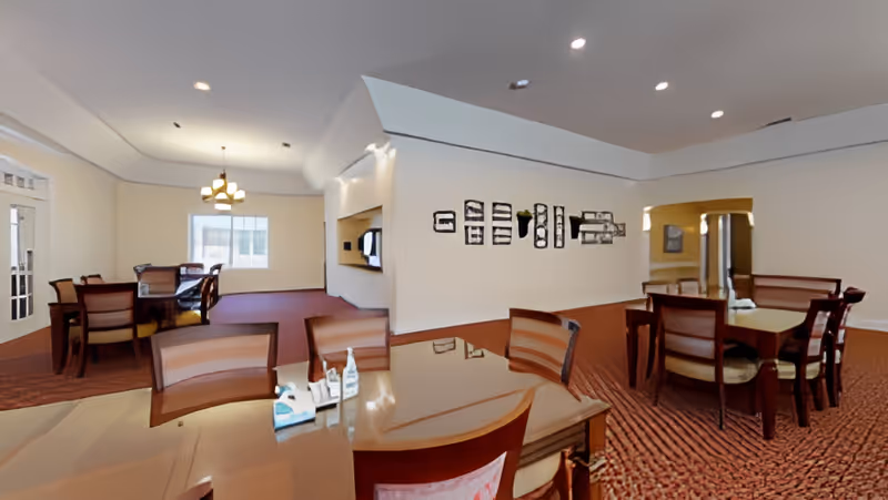 Spacious communal dining room with multiple wooden tables and chairs, carpeted floor, and overhead lighting.