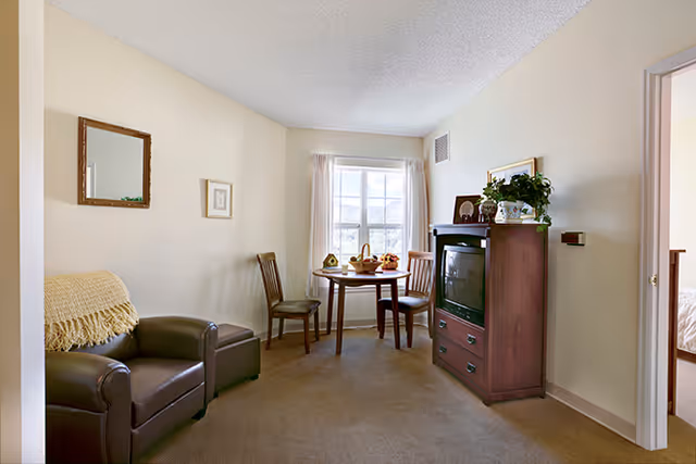 A cozy living area in an assisted living facility featuring a brown leather armchair with a beige throw, a small round wooden table with two chairs near a window with white curtains, and a wooden cabinet with a TV and decorative plants on top. A doorway leads to a bedroom with a visible bed.