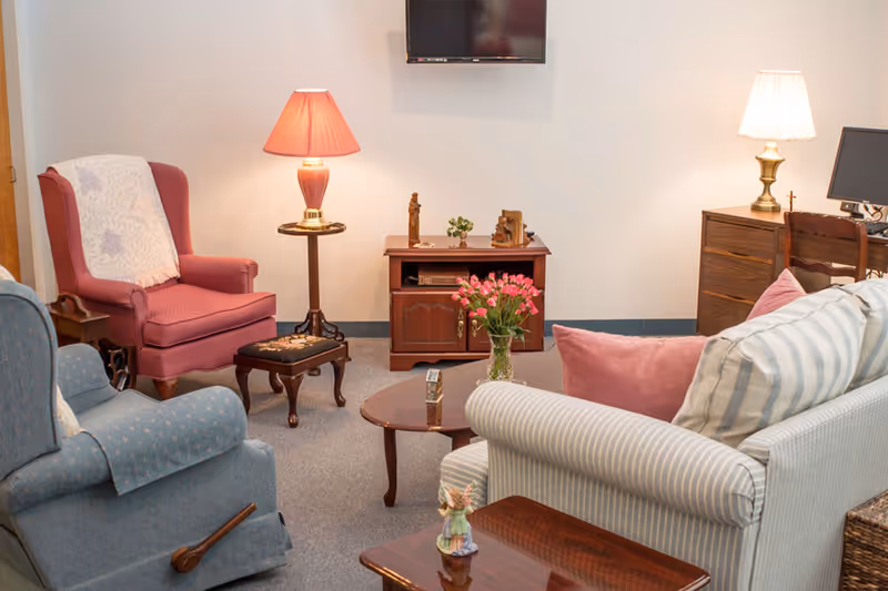 A cozy living room area with a striped sofa adorned with pink pillows, a blue armchair, and a red armchair with a white throw. There are two table lamps on either side, a small wooden coffee table with a vase of pink flowers, a wooden cabinet with decorative items, and a wall-mounted TV above the cabinet.