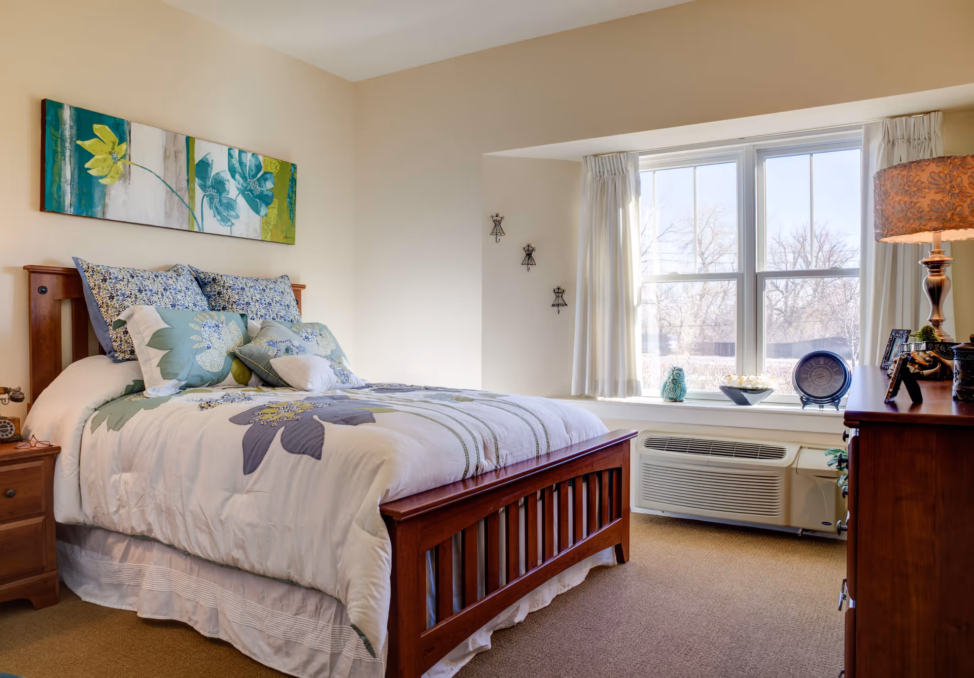 Sunlit bedroom with a wooden bed dressed in floral bedding, bedside tables, a dresser, and a large window with curtains.