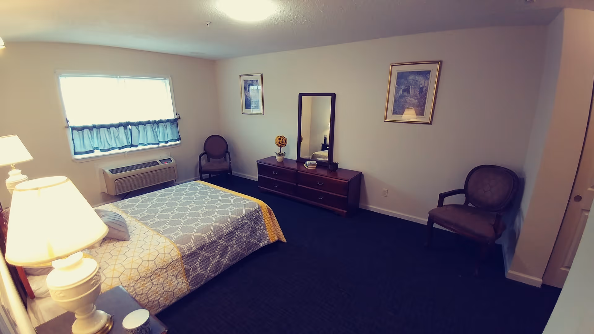 A simple bedroom with a bed covered in a patterned bedspread, two chairs, a dresser with a mirror, a window with a blue curtain, and two framed pictures on the walls. The room has carpeted flooring and soft lighting from a bedside lamp.