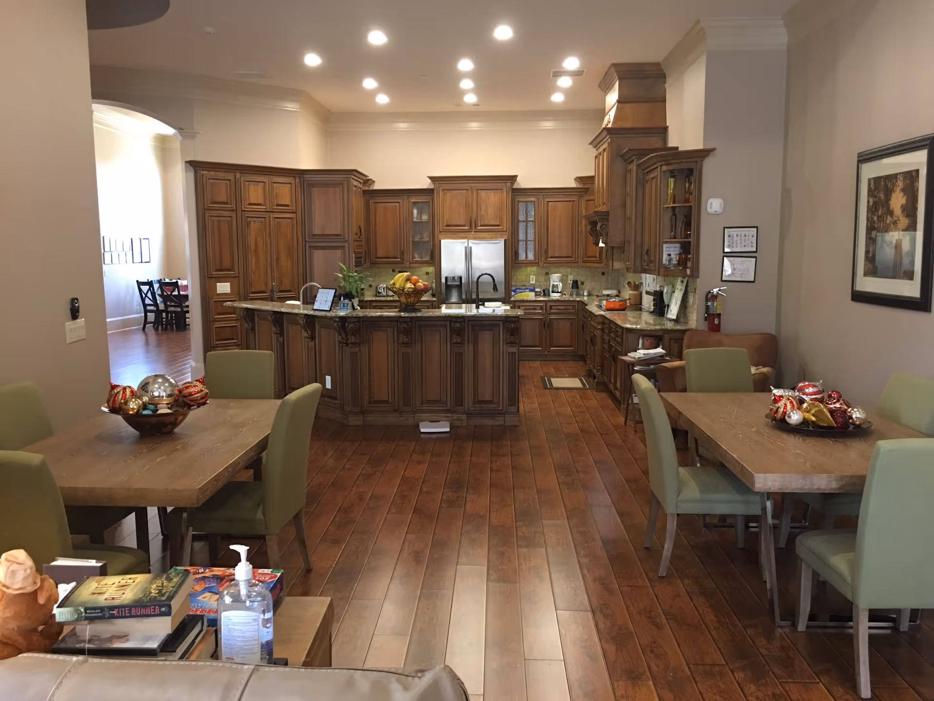 Interior view of a senior living facility kitchen and dining area featuring wooden cabinets, a kitchen island with a sink, stainless steel refrigerator, and two dining tables with green chairs. The floor is wooden, and there are decorative bowls with ornaments on the tables. A hand sanitizer bottle and some books are visible on a side table in the foreground.