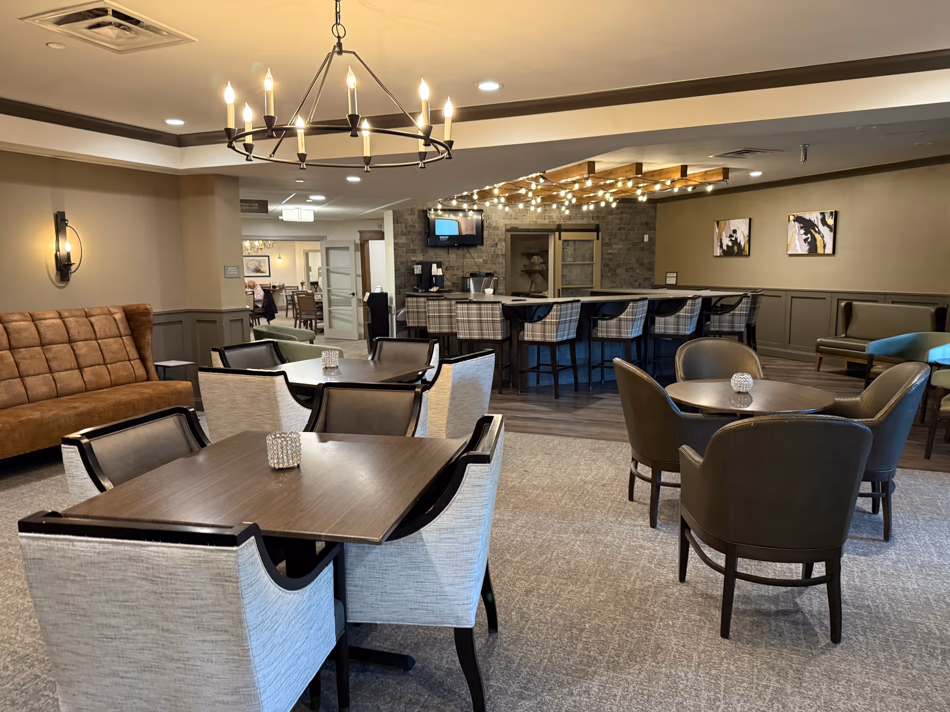 A cozy and well-lit common area in Monarch Bella Terra featuring multiple seating arrangements including tables with chairs, a brown cushioned bench, and a bar area with high stools. The room has warm beige walls, a chandelier, wall sconces, and decorative artwork on the walls.