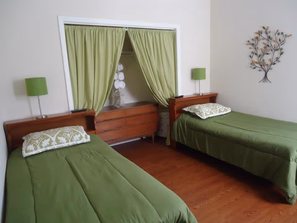 Two twin beds with green bedding and matching lamps in a neatly decorated bedroom with a dresser and green curtains in a recessed closet.