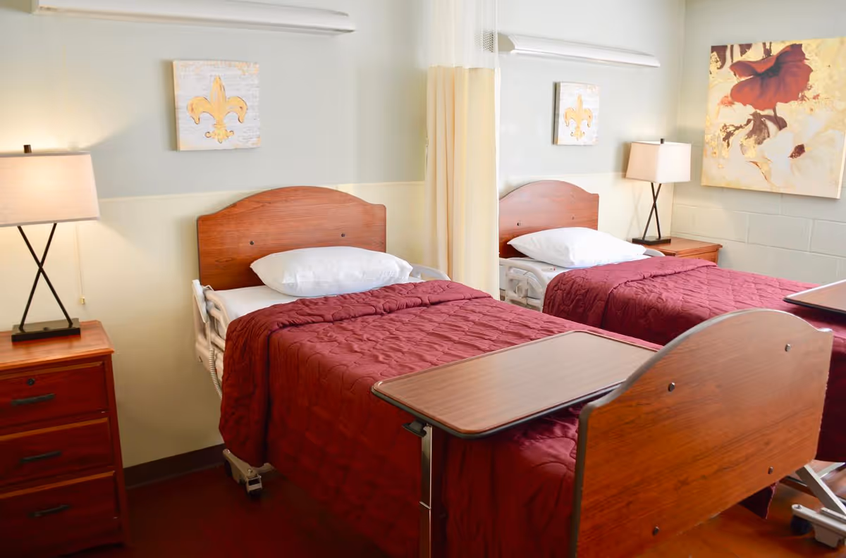 A senior living facility bedroom with two hospital-style beds, each with a wooden headboard and footboard, white pillows, and maroon quilted blankets. Between the beds is a wooden nightstand with a lamp. A beige privacy curtain hangs between the beds. The walls are light-colored with two pieces of wall art, one featuring a fleur-de-lis and the other a floral design.
