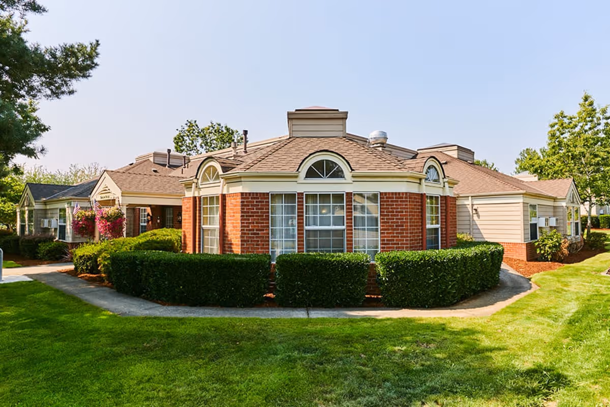 Exterior view of a single-story senior living facility building with a combination of brick and siding walls, multiple windows, and a well-maintained lawn with bushes and trees under a clear sky.