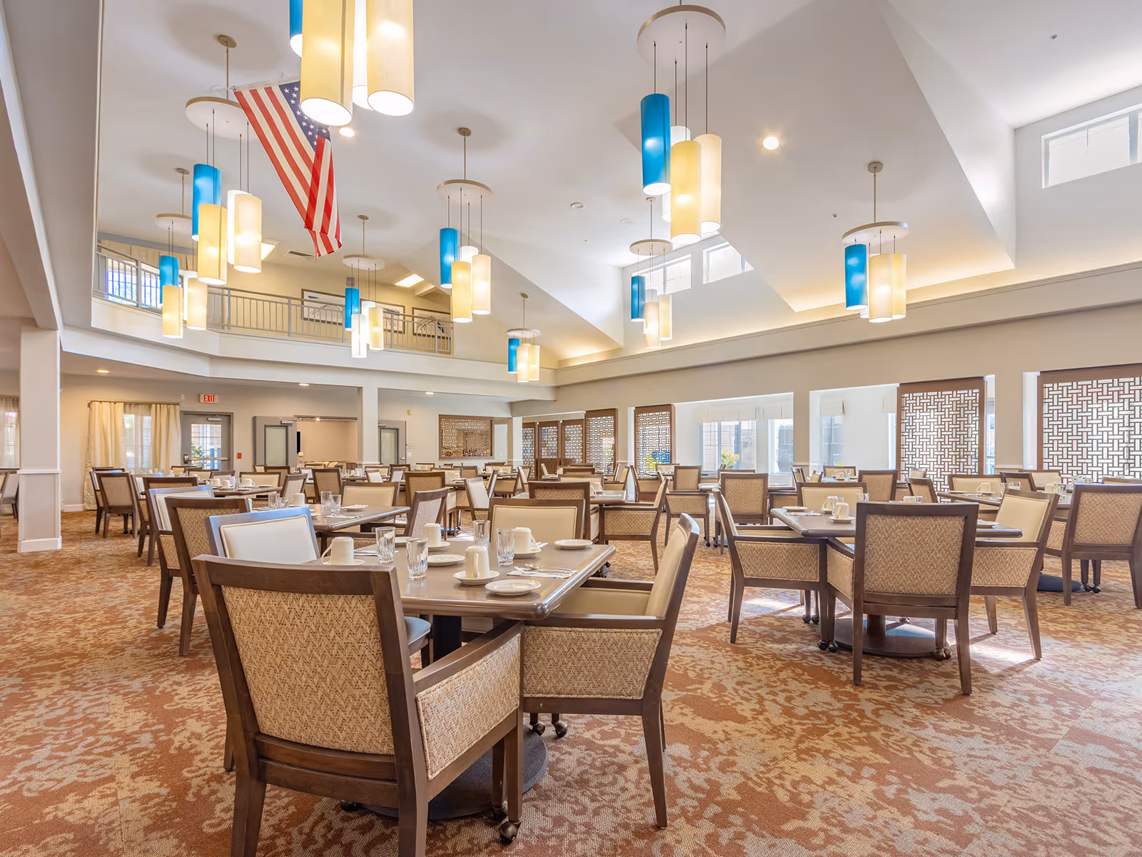 A spacious dining room with multiple tables and chairs arranged neatly. Each table is set with cups, glasses, and plates. The room features high ceilings with modern pendant lights in blue and cream colors. An American flag hangs from the ceiling. Large windows with decorative screens allow natural light to fill the room.