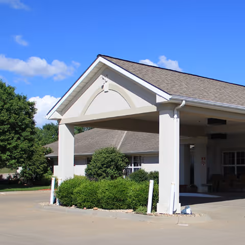 Entrance of a senior living facility with a covered drop-off area supported by white columns, surrounded by green bushes and trees under a clear blue sky.
