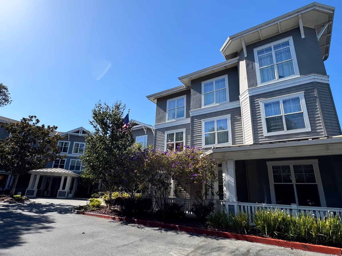 Front exterior of a multi-story gray senior living building with windows, landscaping, and an entrance canopy under a clear blue sky.