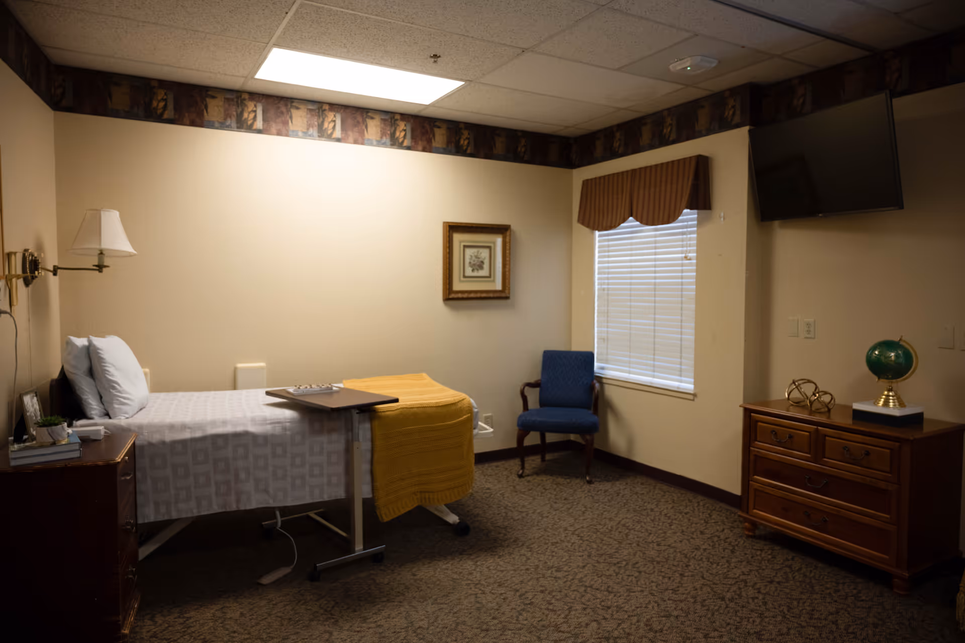 A small, neatly arranged bedroom in a healthcare facility featuring a single bed with white sheets and a yellow blanket, a bedside table with a lamp and some books, a blue upholstered chair near a window with blinds and a brown valance, a wooden dresser with decorative items including a globe, and a wall-mounted TV.