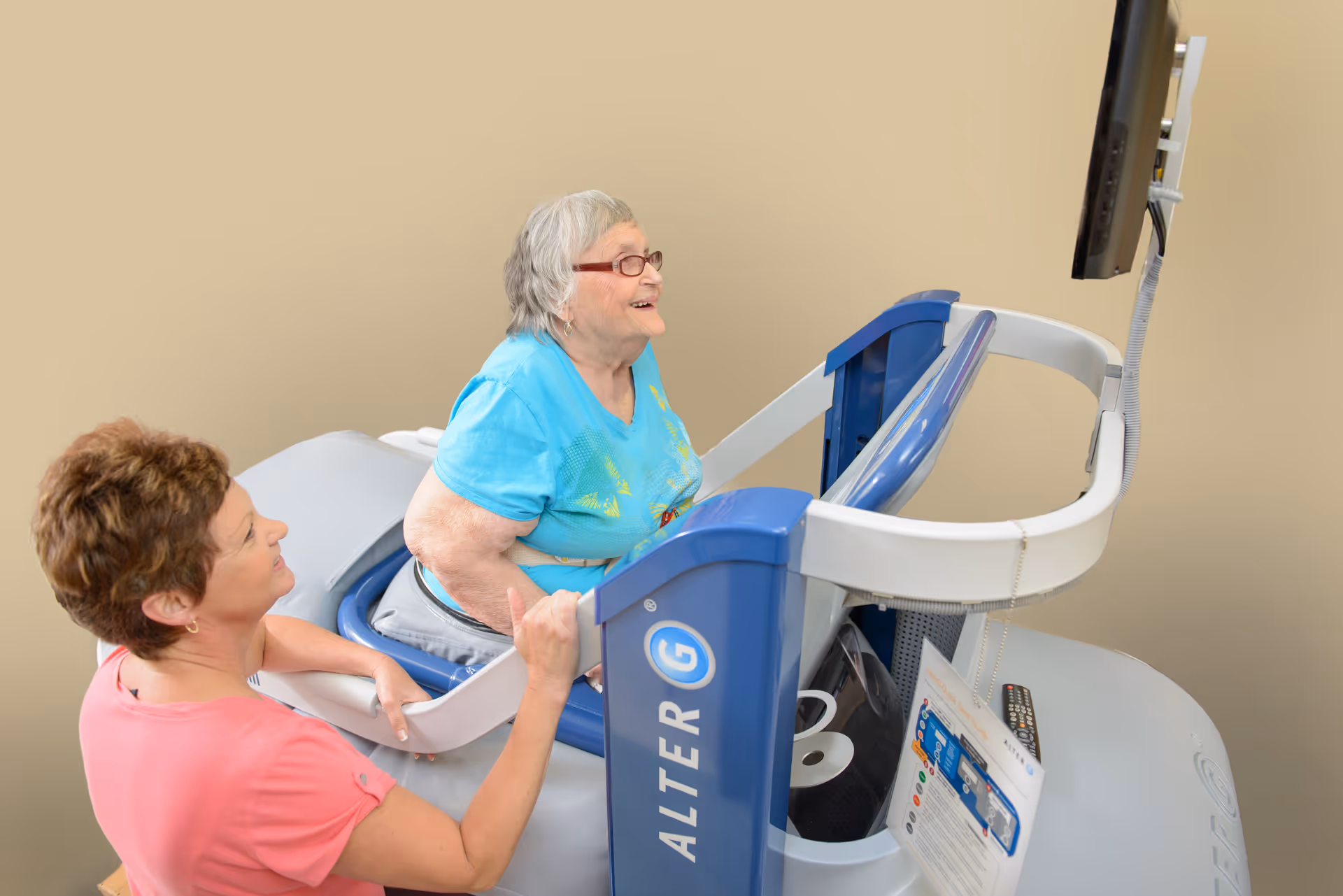 An elderly woman in a blue shirt is using an AlterG anti-gravity treadmill with the assistance of a caregiver in a pink shirt. The woman is smiling and looking at a screen attached to the treadmill.