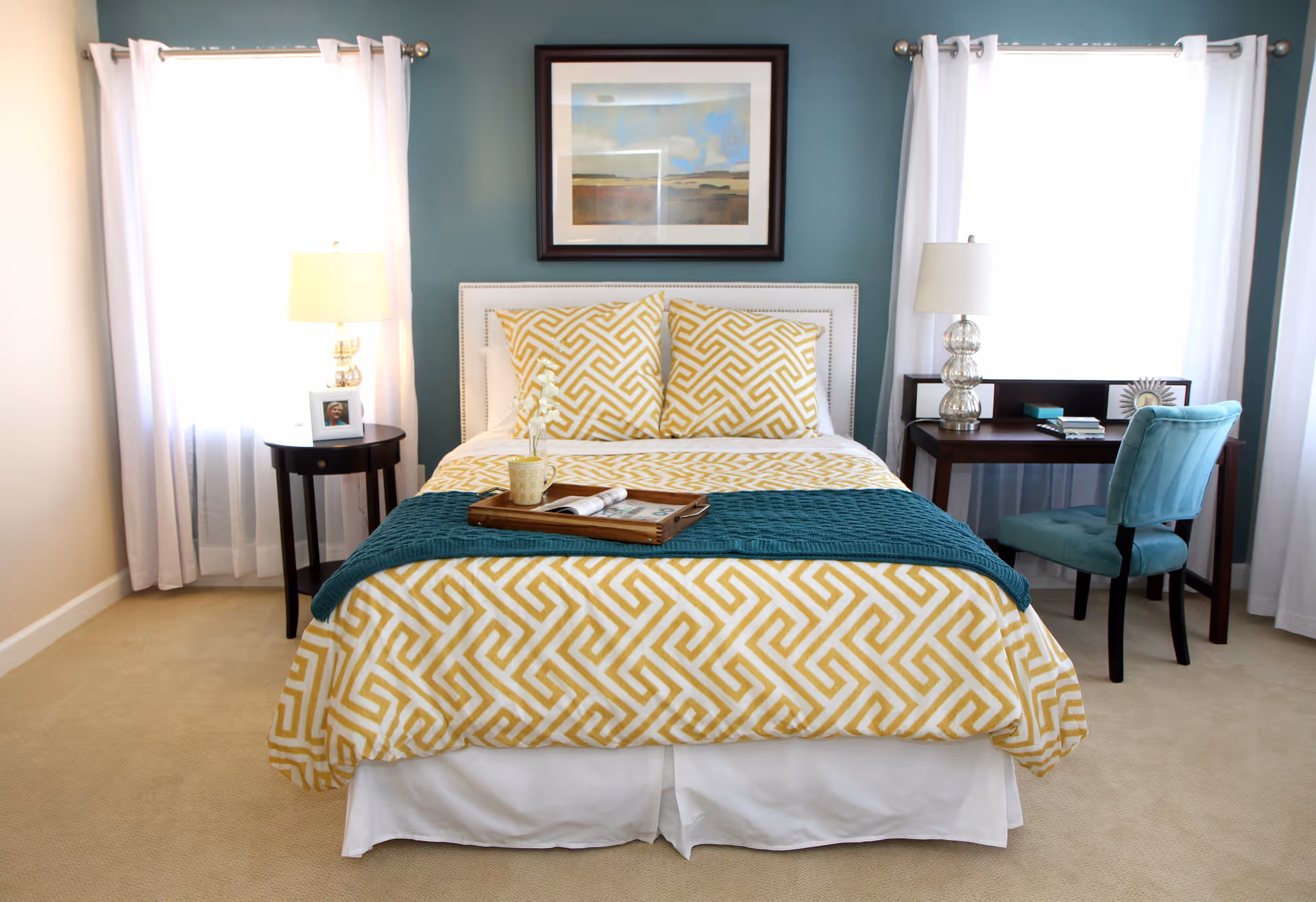A neatly made bed with yellow and white geometric patterned bedding and two matching pillows, a teal throw blanket at the foot of the bed, and a wooden tray with a cup and magazine on it. The bed is flanked by two windows with white curtains, a small round side table with a lamp and framed photo on the left, and a dark wooden desk with a lamp, books, and a teal upholstered chair on the right. A framed landscape painting hangs on the teal accent wall behind the bed.