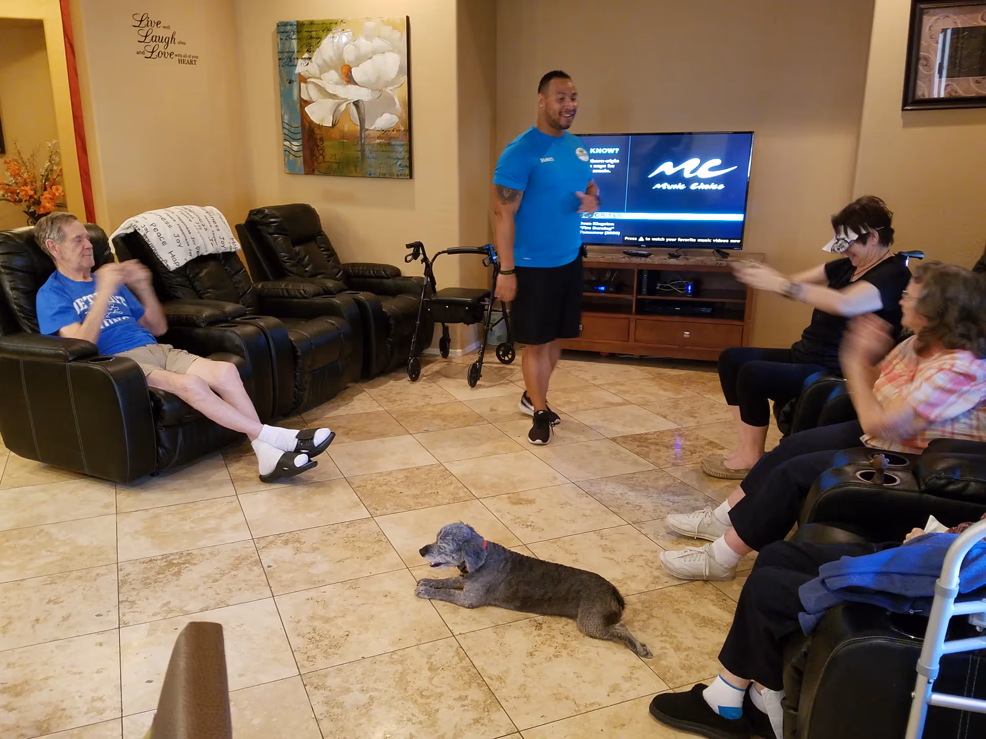 A group of elderly people sitting in a living room with leather recliners, clapping and engaging with a standing man in a blue shirt. A small dog lies on the tiled floor. The room has a TV on a wooden stand, a walker, a floral painting on the wall, and a wall decal that reads 'Live well Laugh often and Love with all of your HEART.'