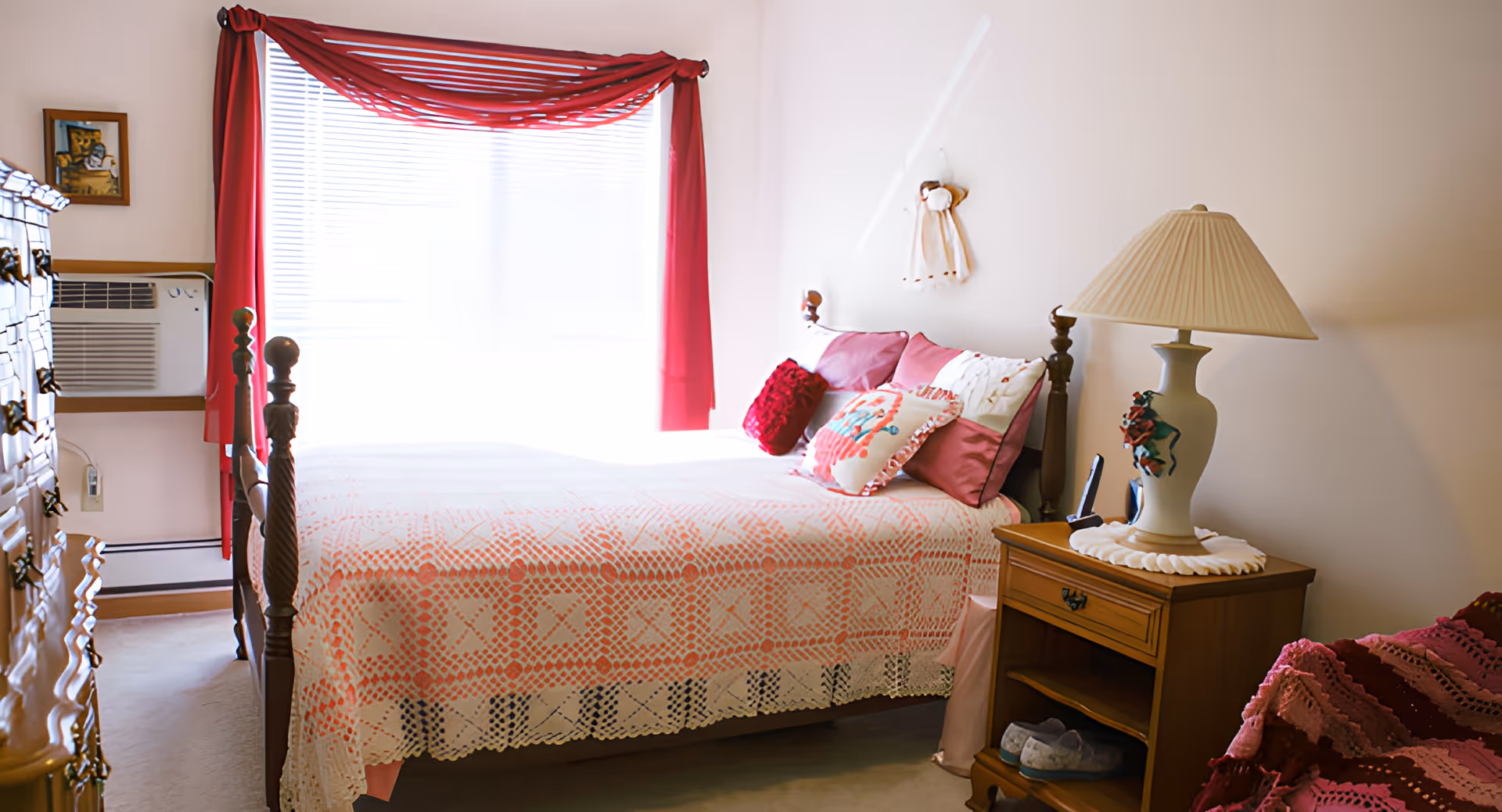 Sunlit bedroom with a neatly made bed covered by a pink patterned quilt, bedside table with a lamp, and red curtains over a window.