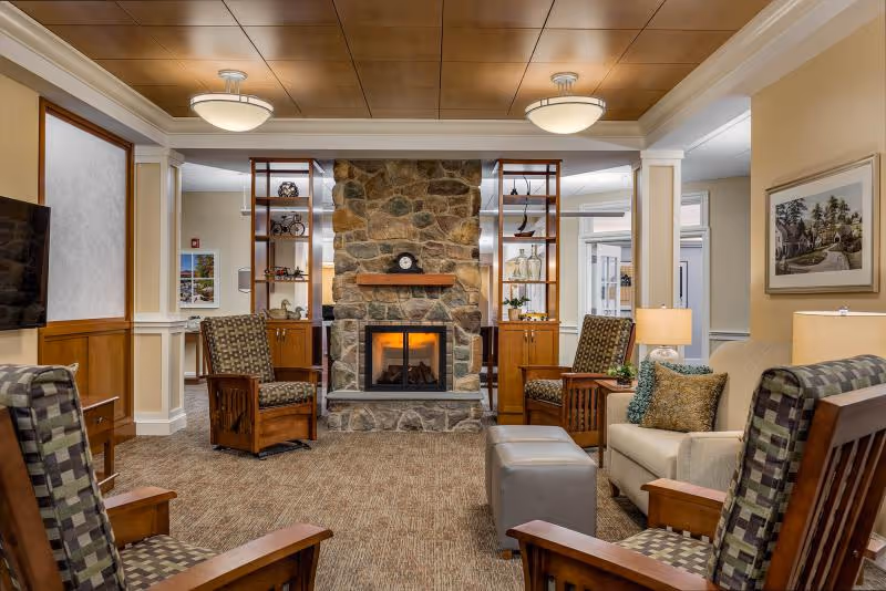 A cozy living room area in a senior living facility featuring a stone fireplace with a wooden mantel, surrounded by wooden shelving units with decorative items. The room has several cushioned armchairs with patterned upholstery, a beige sofa with decorative pillows, two ottomans, a carpeted floor, and warm lighting from ceiling fixtures and table lamps. A framed picture hangs on the wall to the right.