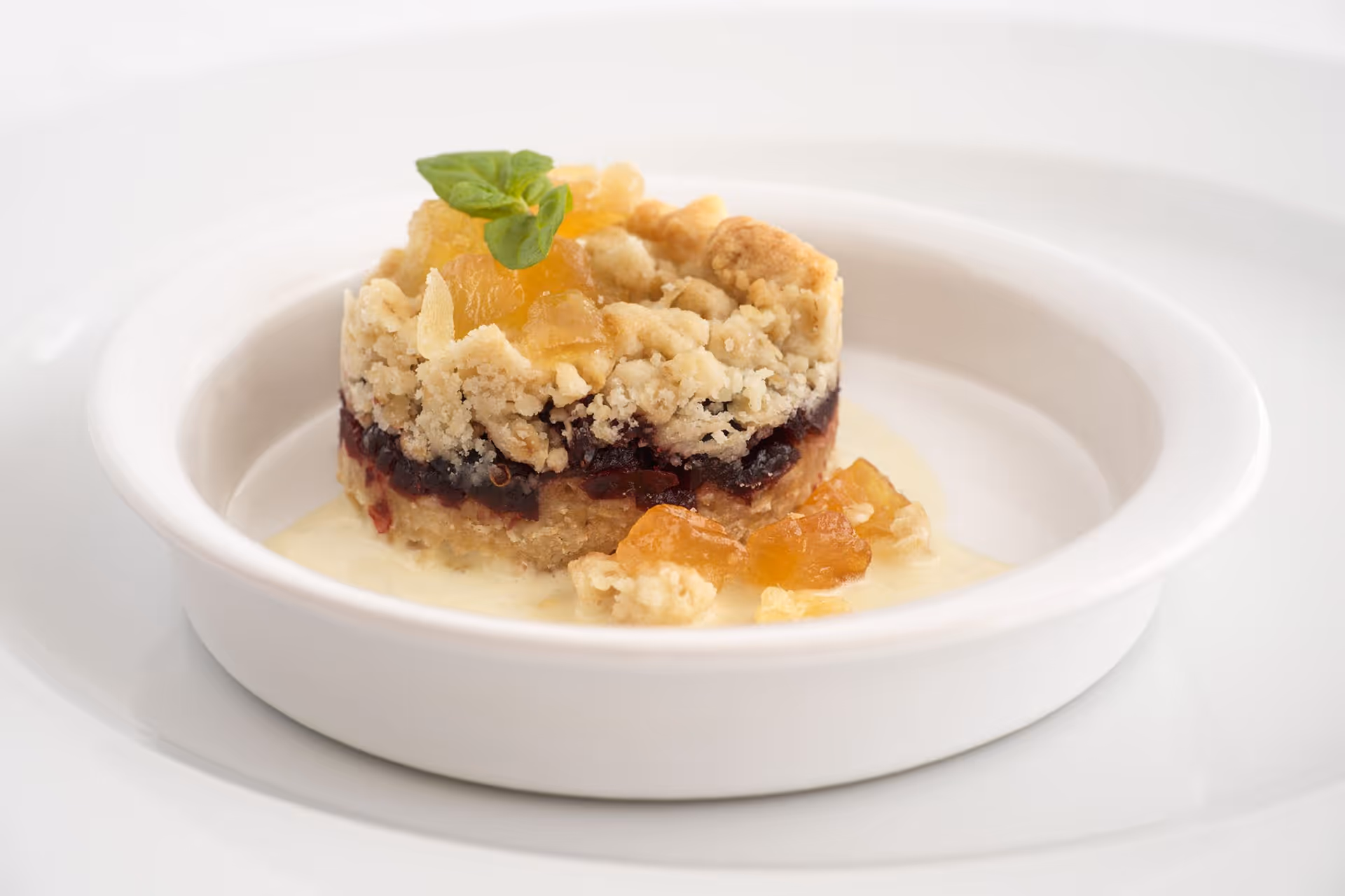 A close-up of a layered dessert served in a white bowl, featuring a crumbly topping, a dark fruit layer, and garnished with a small green leaf and pieces of fruit on a creamy sauce.