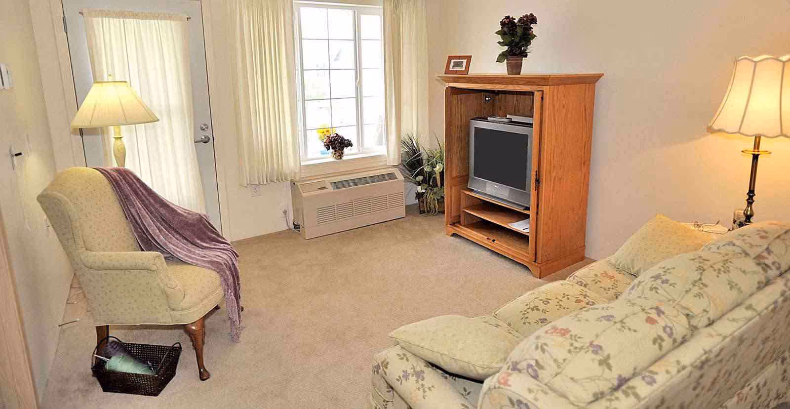 A cozy living room with a floral patterned sofa, an armchair draped with a purple throw, a wooden TV cabinet with a television, a window with cream-colored curtains, and two lamps providing warm lighting.