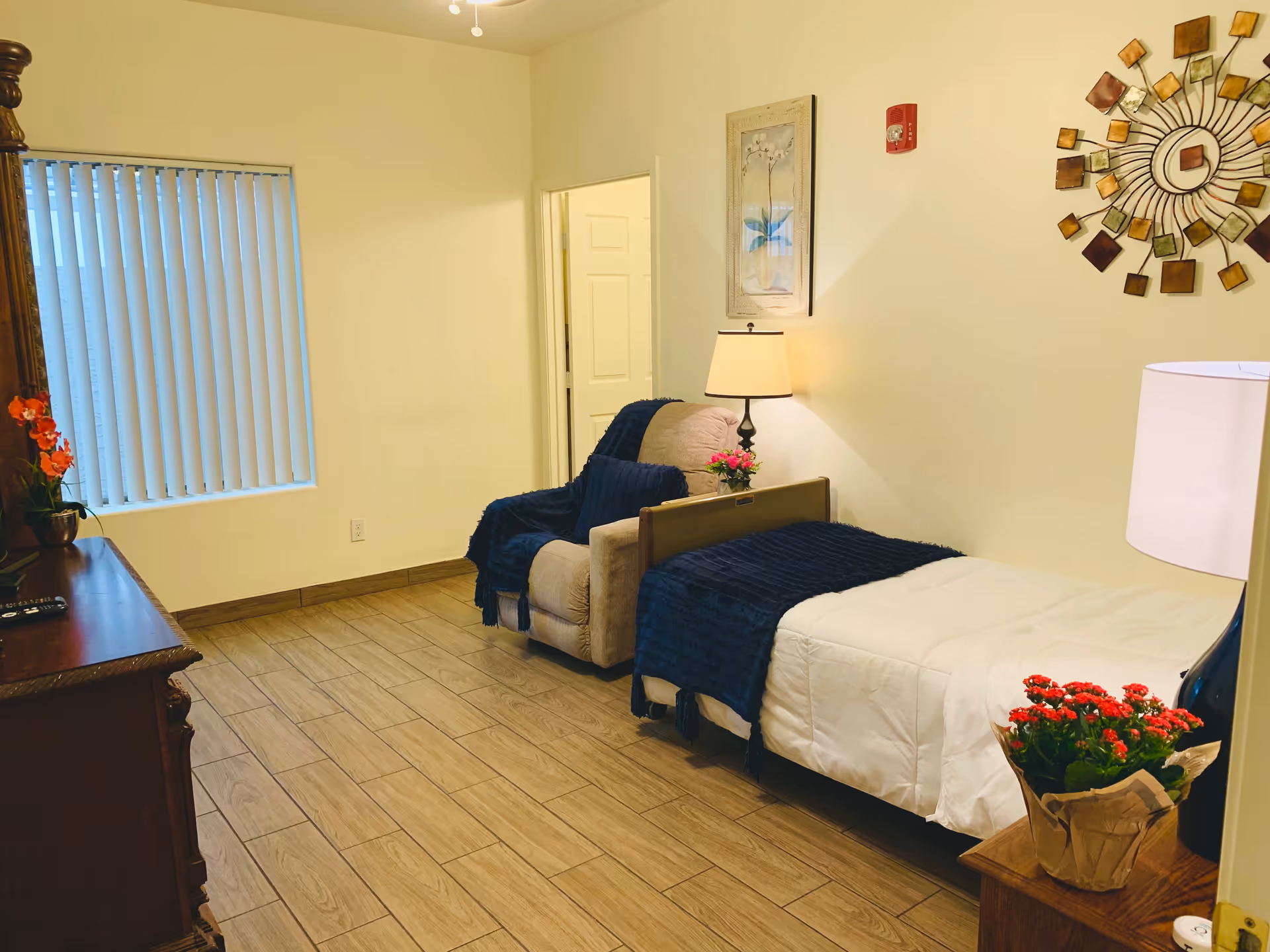 A tidy bedroom with a single bed, upholstered armchair, lamps, dresser, wall art, and potted flowers.