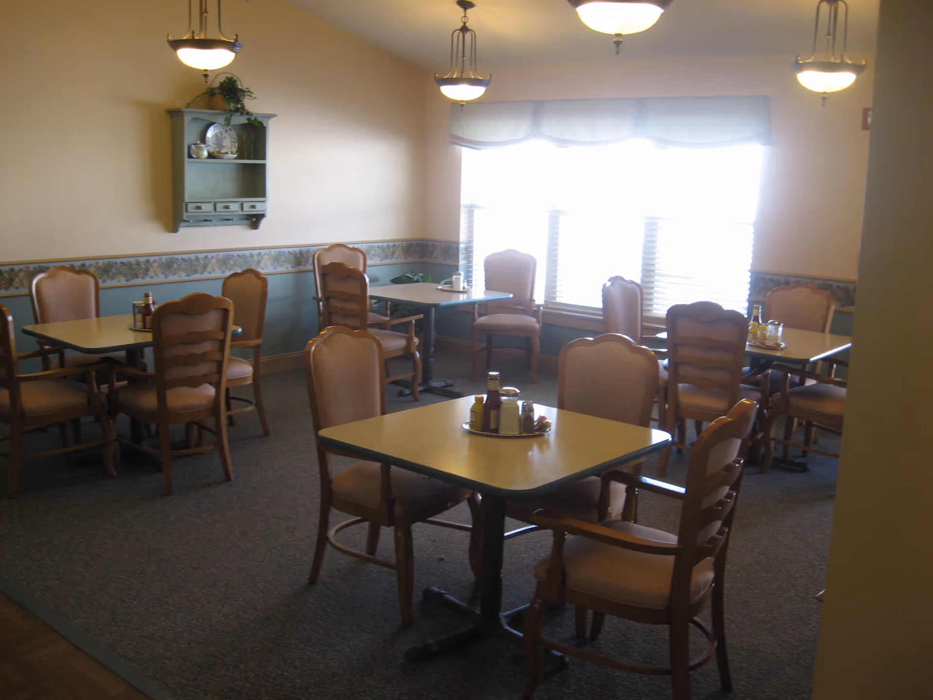 A dining room with several square tables and wooden chairs with cushioned seats and backs. The walls are painted beige with a floral wallpaper border and a green wainscoting. There are three hanging light fixtures on the ceiling and a window with blinds and a valance letting in natural light. A small wall shelf with decorative items is mounted on one wall.