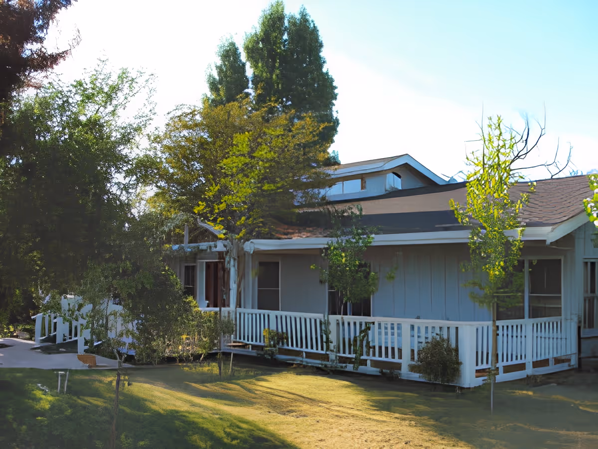 A single-story residential building with a white exterior and a porch surrounded by a white railing. The building is partially shaded by several trees and bushes, with a well-maintained grassy lawn in the foreground under a clear blue sky.