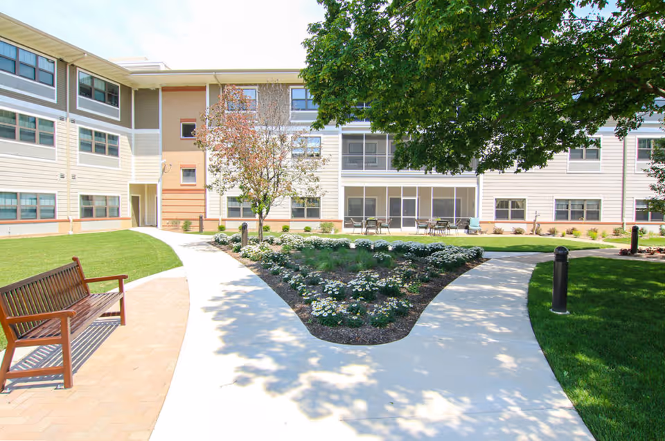 Outdoor courtyard area at EverTrue Lutheran Hillside Village featuring a paved walkway splitting around a flower bed with white flowers and a small tree. There is a wooden bench on the left side and patio tables with chairs near the building. The building has multiple windows and light-colored siding. A large tree provides shade on the right side.