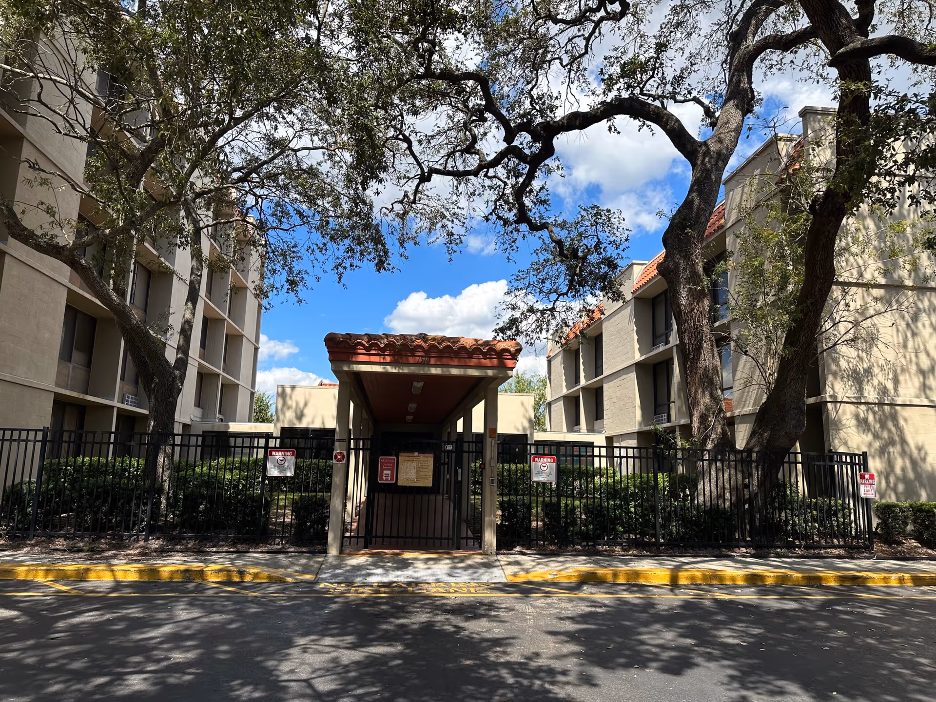 Exterior view of Angels Senior Living at Hacienda Villas showing a gated entrance with a covered walkway. The building has beige walls and red-tiled roofs, with large trees providing shade. The sky is blue with some clouds.