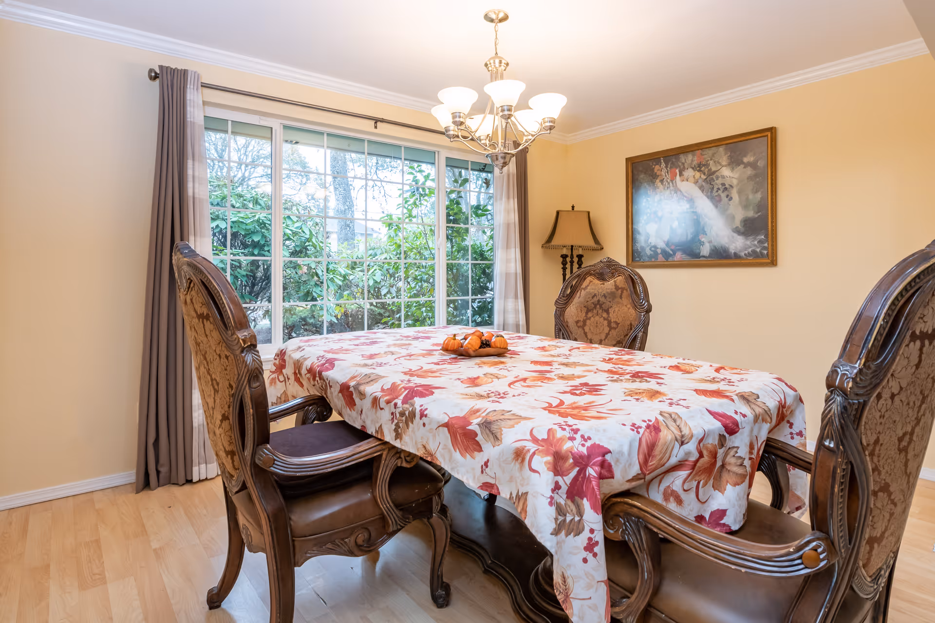 A dining room with a wooden table covered by a floral tablecloth in autumn colors. Four ornate wooden chairs with cushioned seats surround the table. A chandelier with multiple lights hangs above the table. A large window with gray curtains shows greenery outside. A framed painting and a floor lamp are visible on the beige walls.