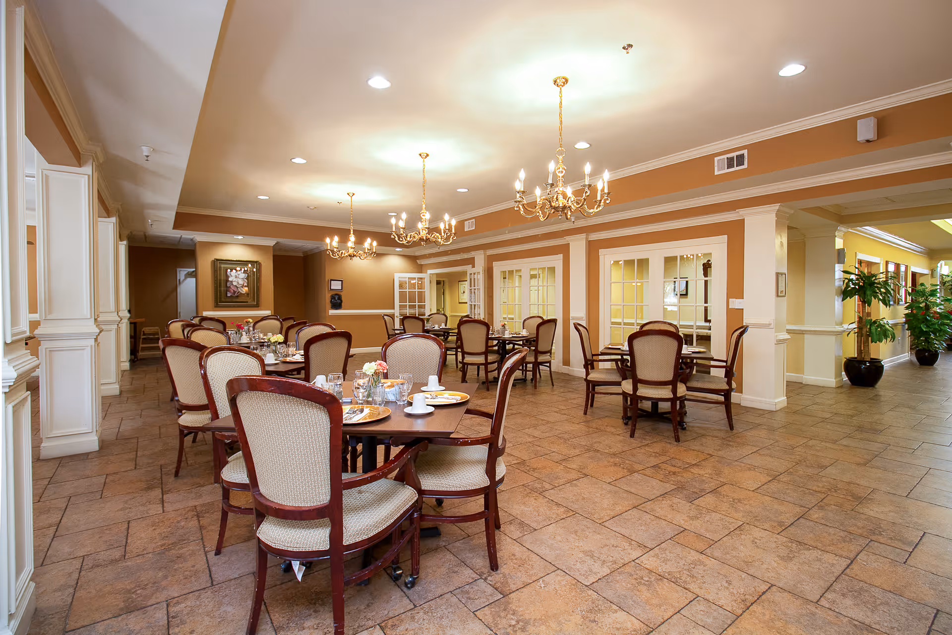 A spacious dining room with multiple round wooden tables and cushioned chairs arranged neatly. The tables are set with plates, cups, and floral centerpieces. The room features warm beige walls, tiled flooring, elegant chandeliers hanging from the ceiling, and large windows with white frames. There are plants and framed artwork on the walls, creating a welcoming atmosphere.