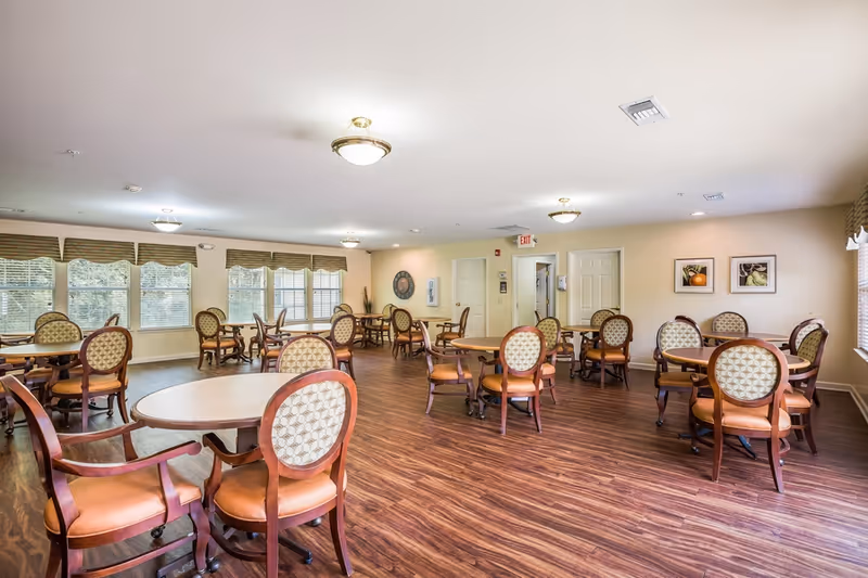 Bright communal dining room with multiple round tables and upholstered chairs on wood flooring.