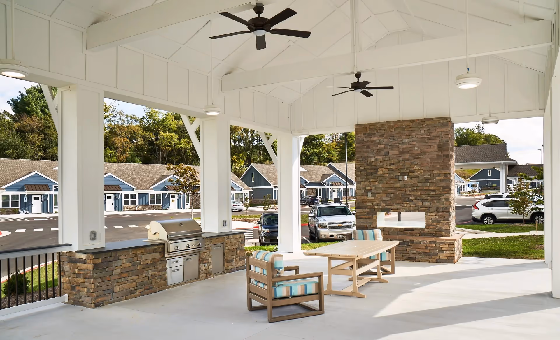 Covered outdoor patio area with a built-in stone grill and countertop, two cushioned chairs with striped upholstery, a wooden table, ceiling fans, and a stone fireplace. In the background, there are blue residential buildings, parked cars, and trees.