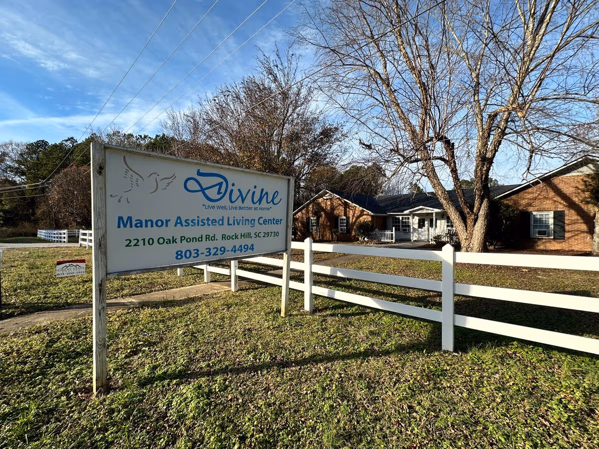 A large sign for Divine Manor Assisted Living Center in front of a single-story brick building with a white fence and leafless trees.