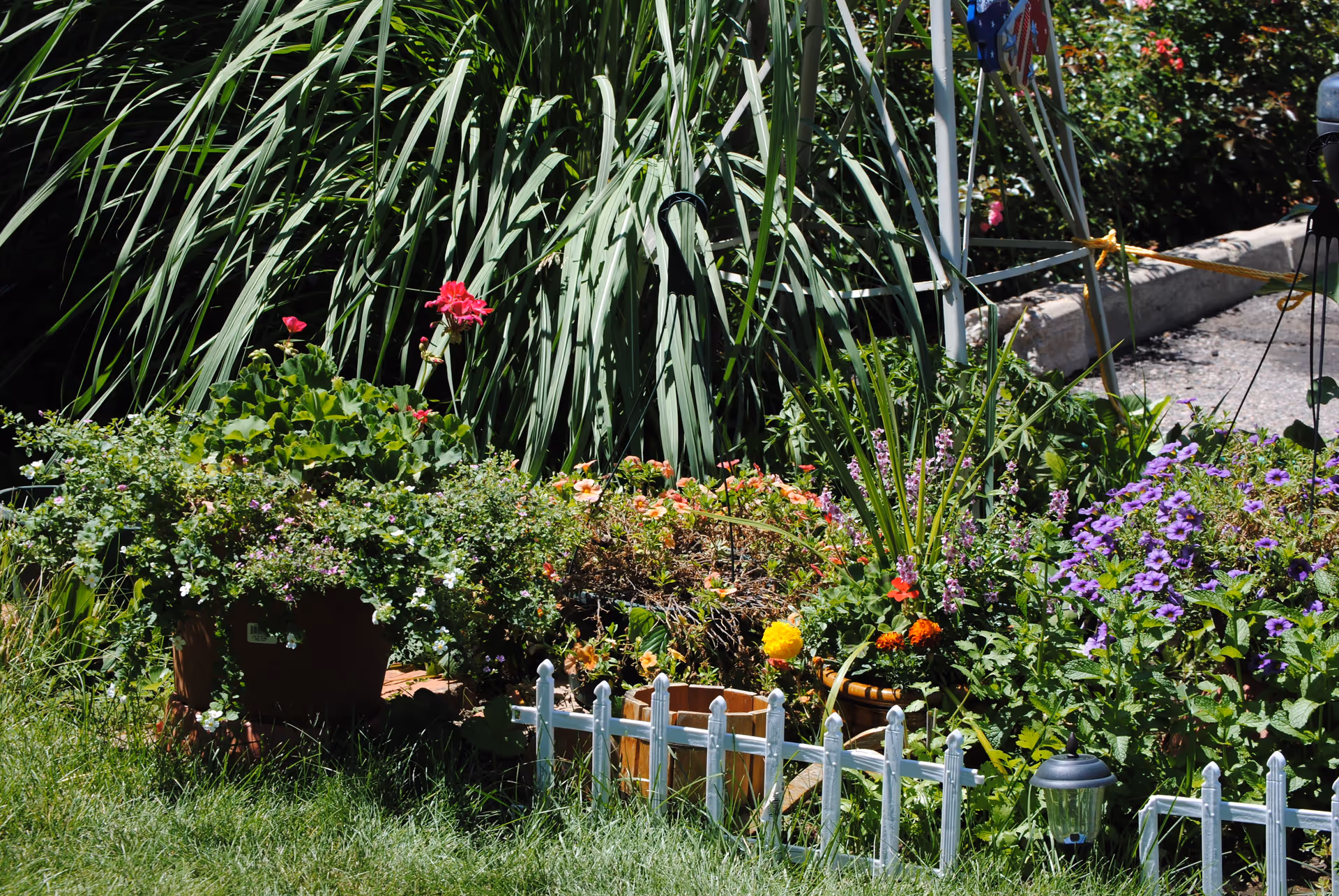 A vibrant garden area with various colorful flowers and green plants, bordered by a small white decorative fence and a grassy lawn in front. There is a paved pathway and some garden decorations visible in the background.