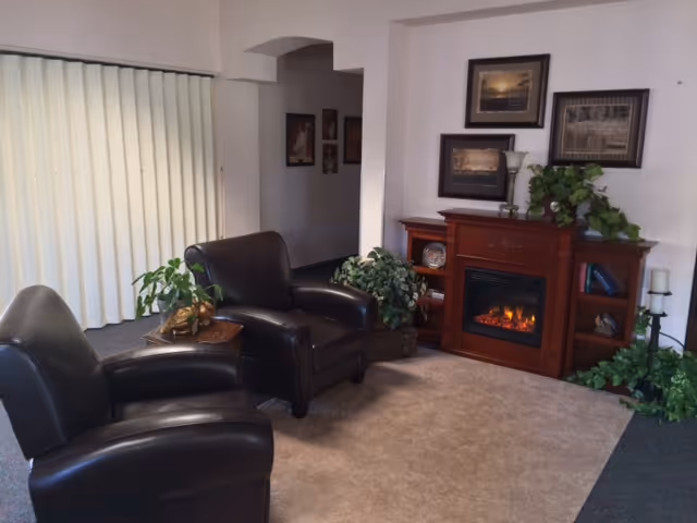 Cozy living room with two dark leather armchairs, a wooden electric fireplace mantel, framed pictures and potted plants.