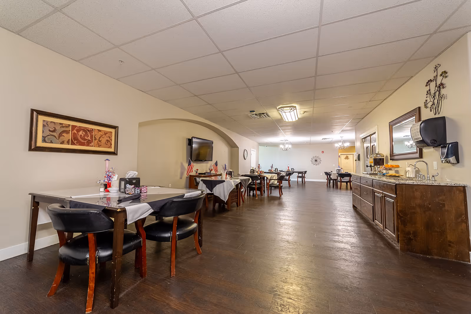 Interior view of a dining room in a senior living facility with several tables and chairs arranged along the walls. The tables are covered with tablecloths and have small decorations. On the right side, there is a long counter with a sink, beverage dispensers, and a mirror above it. The room has a drop ceiling with fluorescent lighting and a wall-mounted TV on the left side.