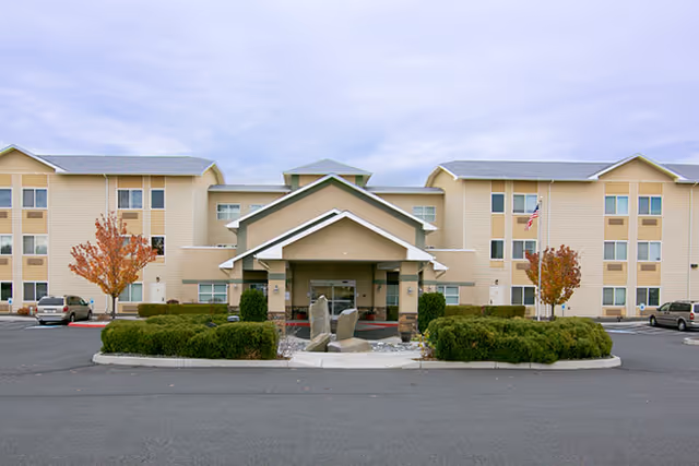 Front exterior view of a three-story senior living facility building with beige siding, a covered entrance, landscaped bushes, and two small trees with autumn foliage. Several cars are parked in the parking lot in front of the building under a cloudy sky.