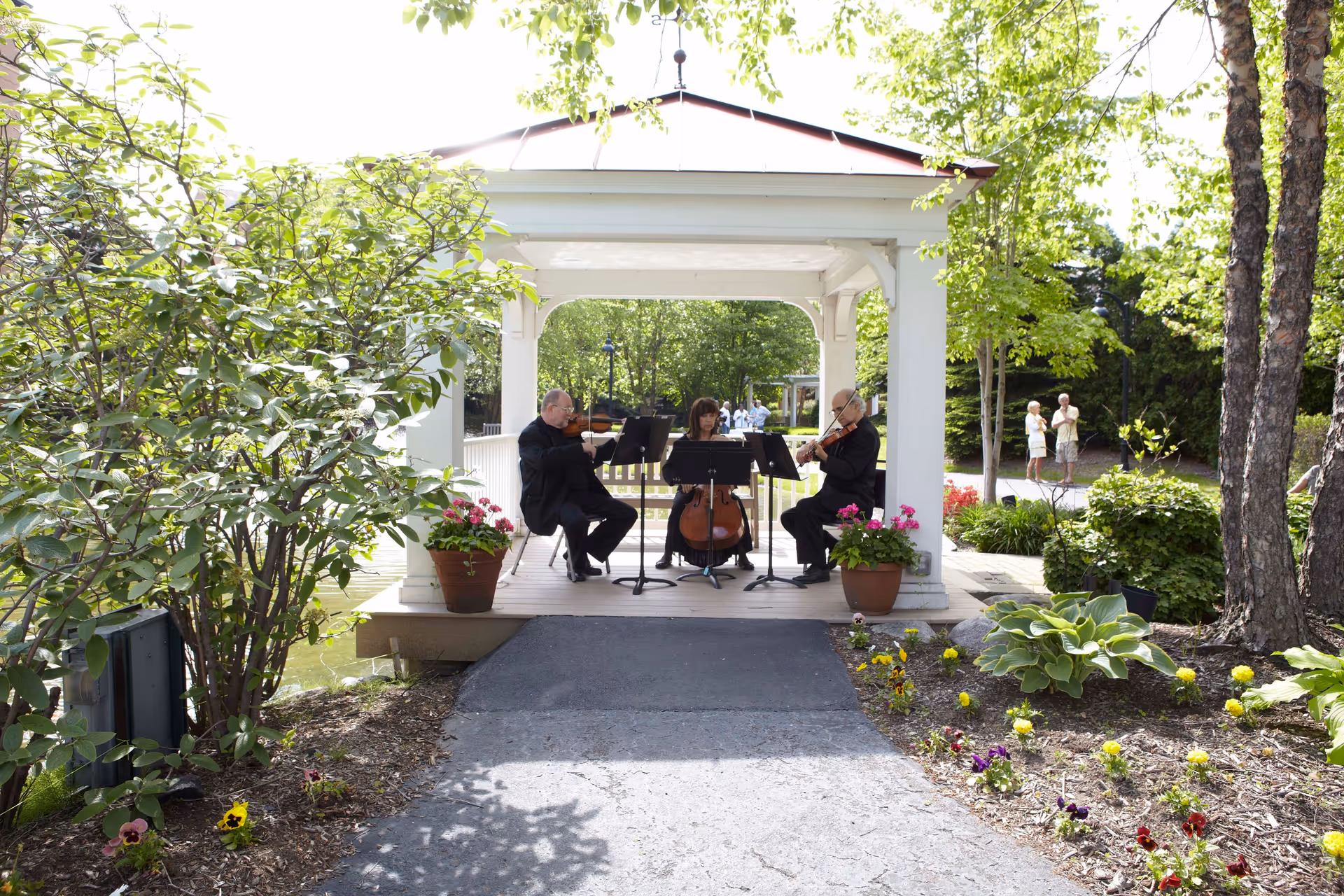 Three musicians playing string instruments under a white gazebo in a garden setting with green trees, flowering plants, and a paved pathway. Two people are standing and talking in the background.