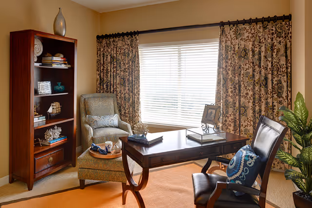 A sunlit sitting room with a wooden desk, leather and upholstered chairs, a bookshelf, and a large window with patterned curtains.