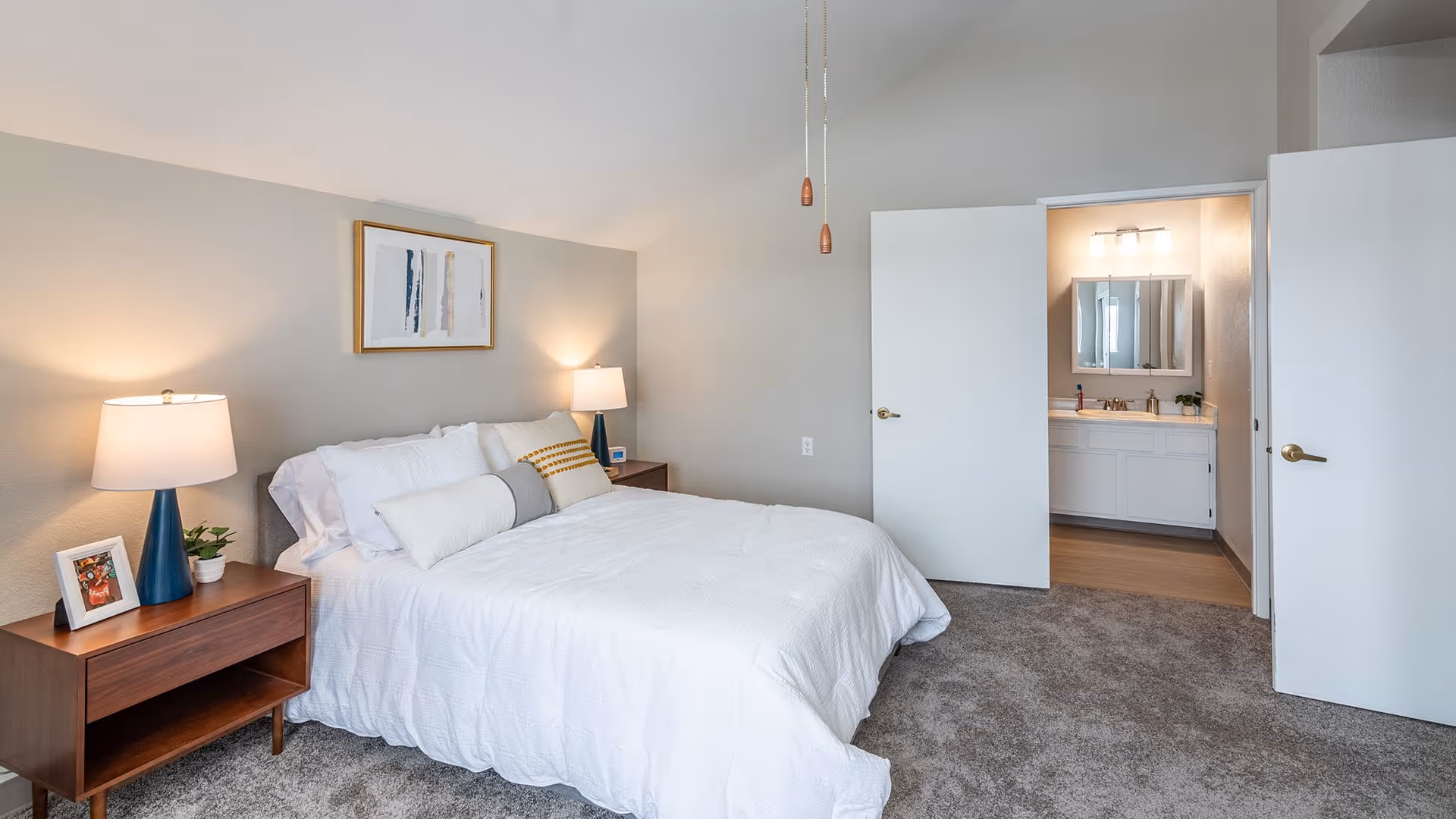 A neatly made bed with white bedding and multiple pillows in a bedroom. There are two wooden nightstands on either side of the bed, each with a lamp and decorative items. The room has beige walls and carpeted flooring. An open door reveals a bathroom with a sink, mirror, and lighting fixture.